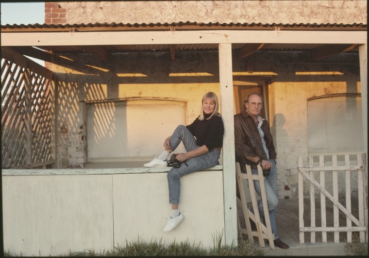 a man and a woman stand in front of an old building as the sun hits them. 