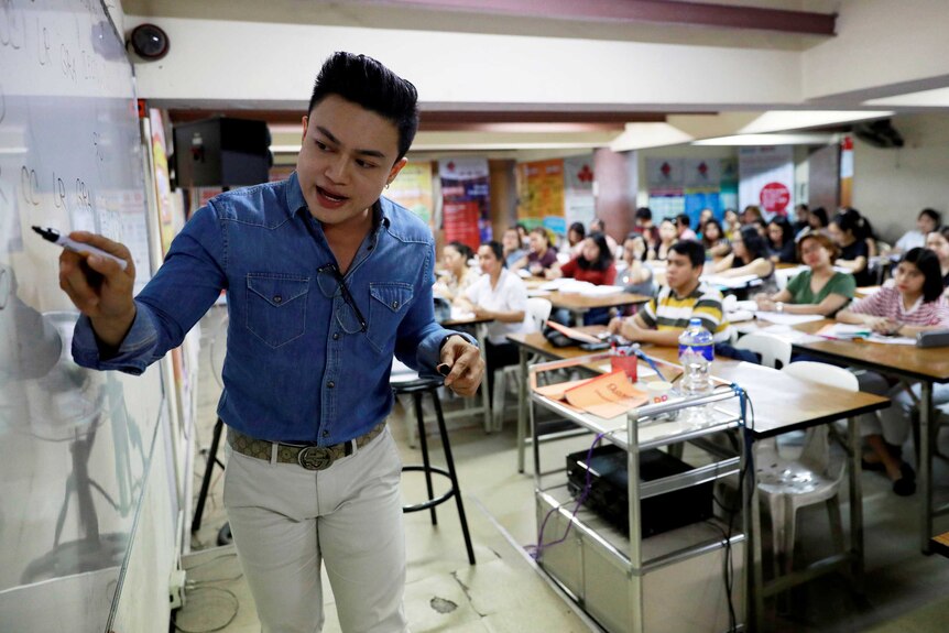 A lecturer in a denim shirt and white jeans points with a marker on a whiteboard while rows of students look behind him.