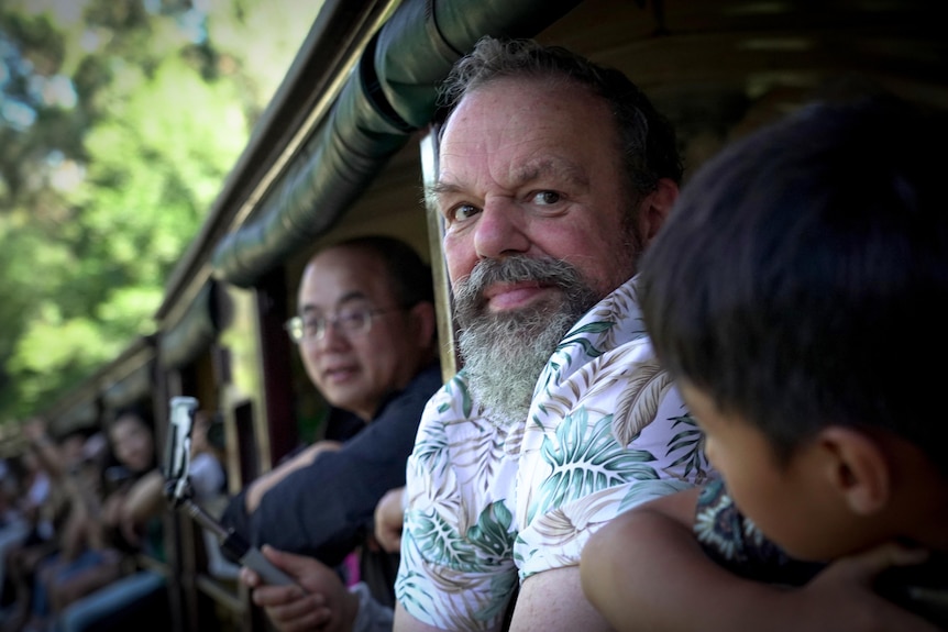 A man with a beard sits leaning out a steam train surrounded by other passengers