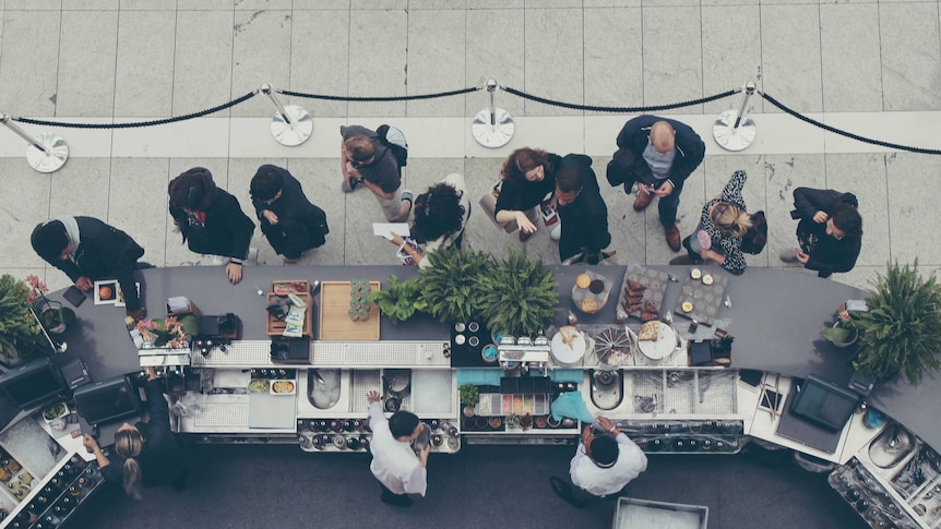 An aerial shot of a queue of people in front of an open-air cafe.