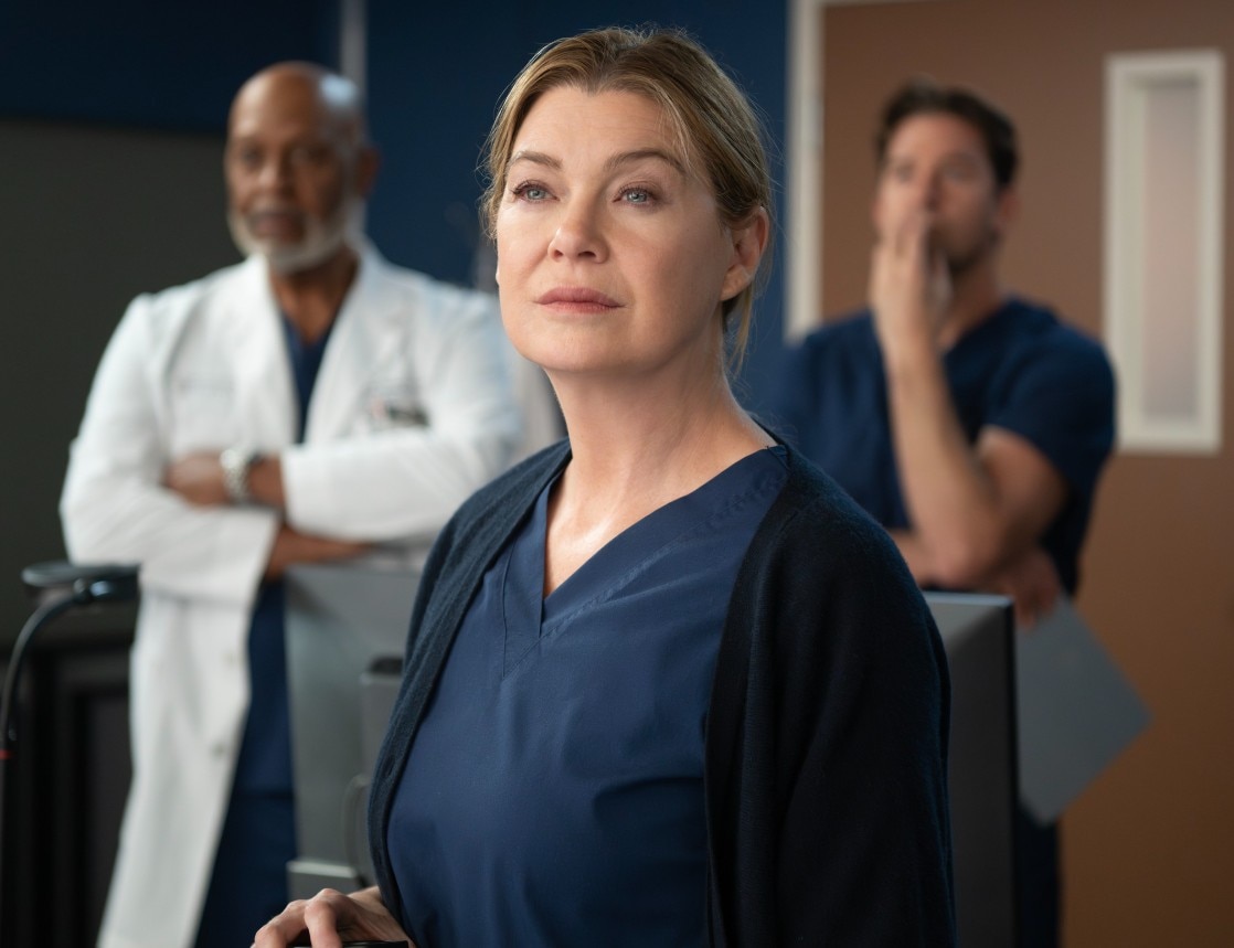 Una mujer con una bata azul oscuro y un cárdigan negro que parece seria. Al fondo, está flanqueada por dos hombres.