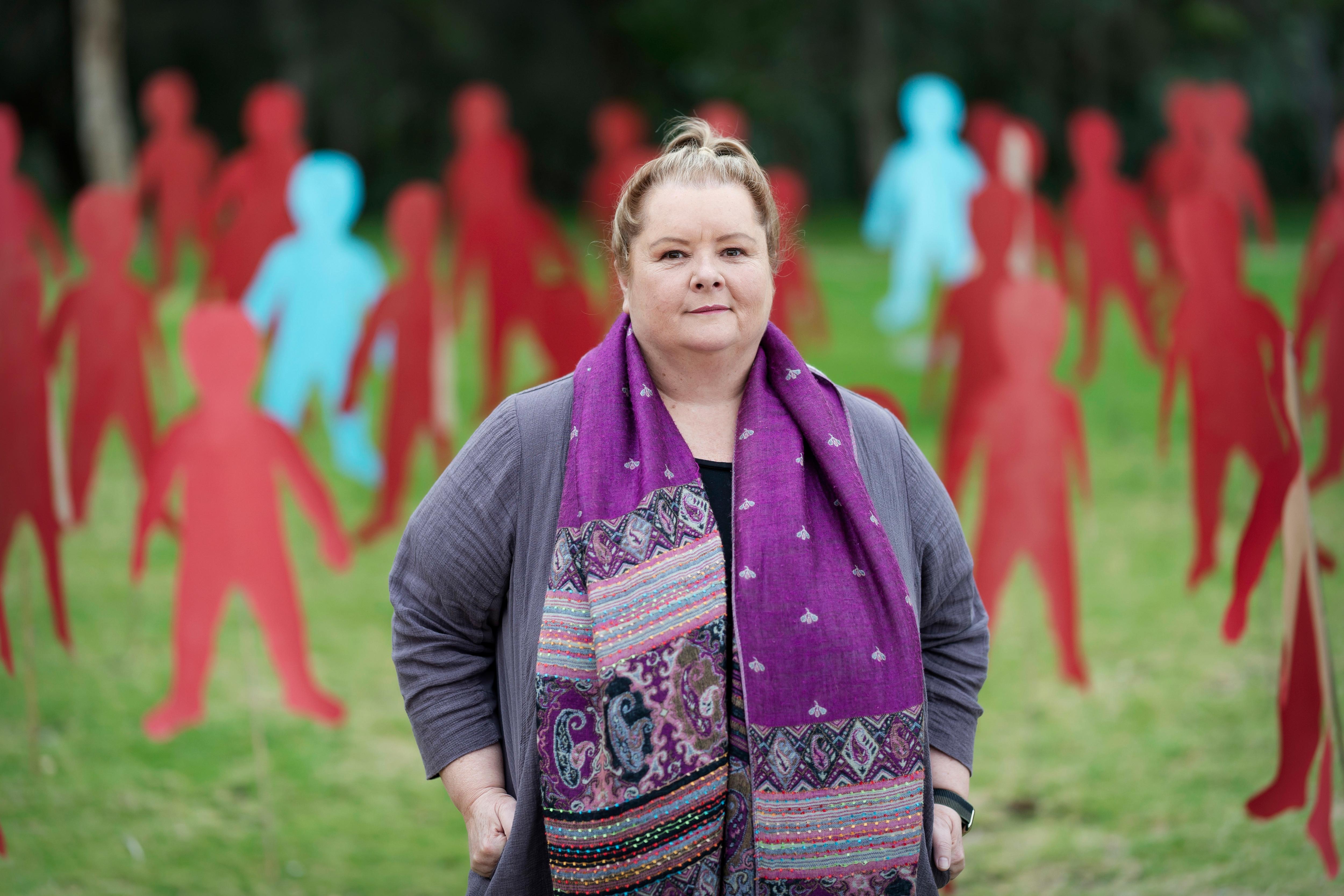 A woman wearing a purple patterned scarf stands in a park in front of cardboard cut out people.