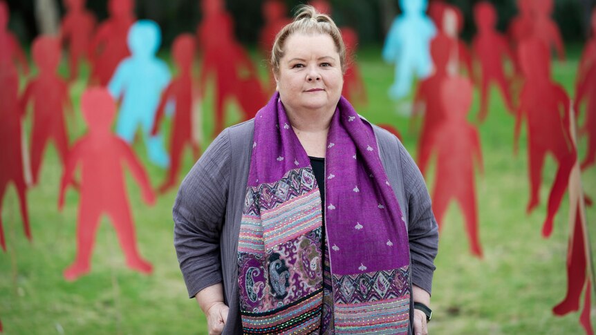 A woman wearing a purple patterned scarf stands in a park in front of cardboard cut out people.