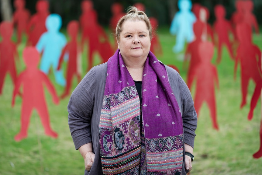 A woman wearing a purple patterned scarf stands in a park in front of cardboard cut out people.