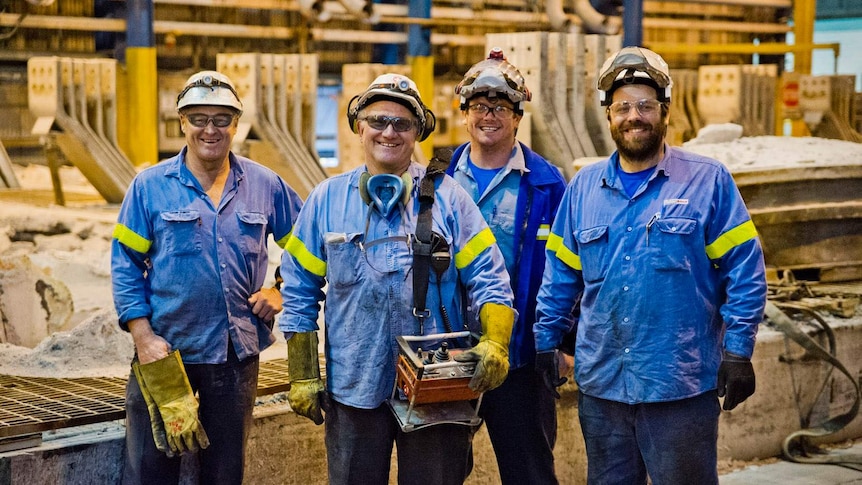 Four men in blue shirts and hard hats standing in part of an aluminium smelter workshop.