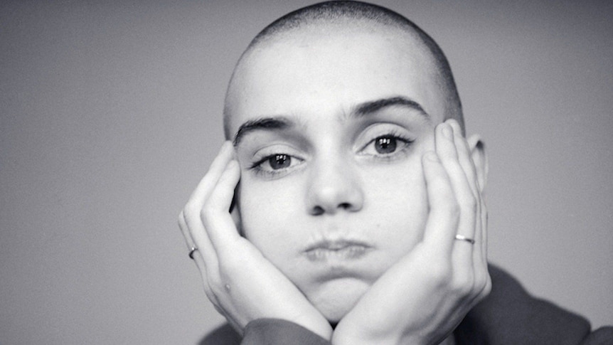 black and white portrait of singer Sinéad O'Connor who has a shaved head and puffs her cheeks out