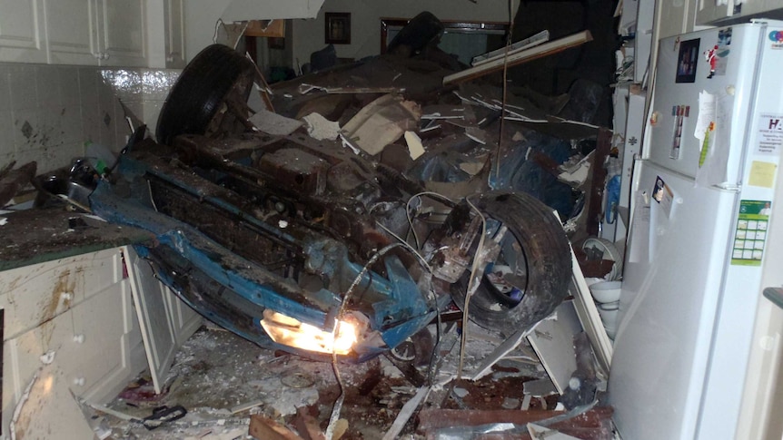 A car lies upside down amidst the wreckage of a kitchen.
