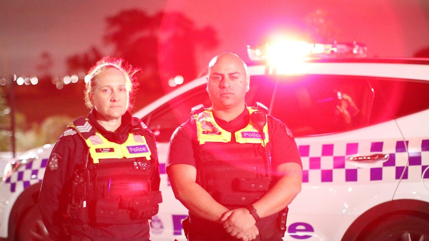 Two police officers, one a woman with her hair tied back and one a man with a bald head, stand in front of their police car.