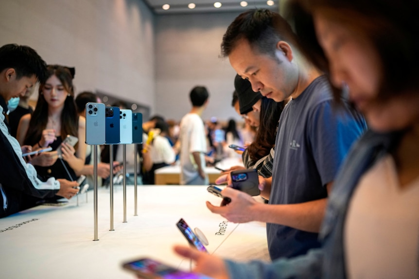 A close up of people standing near an iPhone 15 display in an Apple store, holding the devices as others sit on stands near them