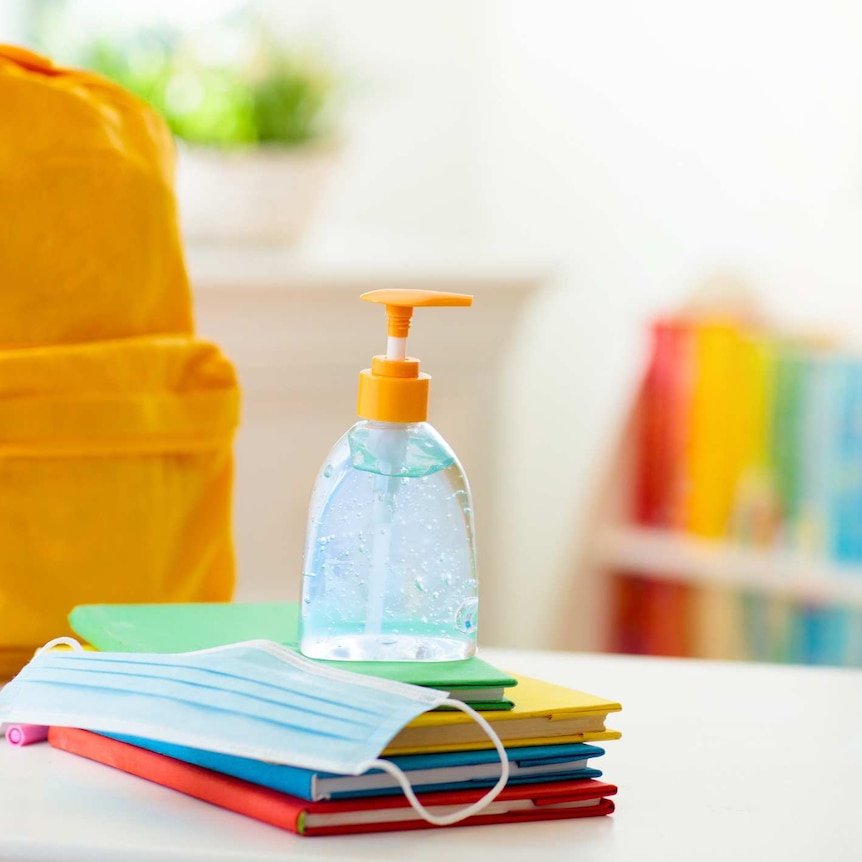 A bright yellow children's bag sits next to a stack of coloured books, hand sanitiser and a facemask in a well-lit classroom.