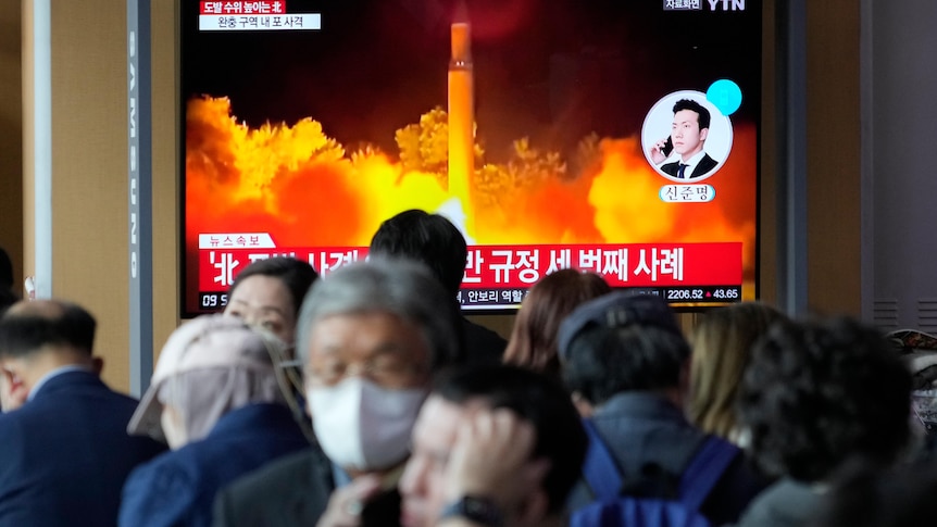 people wearing masks stand in front of a tv screen showing a missile being launched