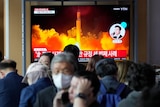people wearing masks stand in front of a tv screen showing a missile being launched