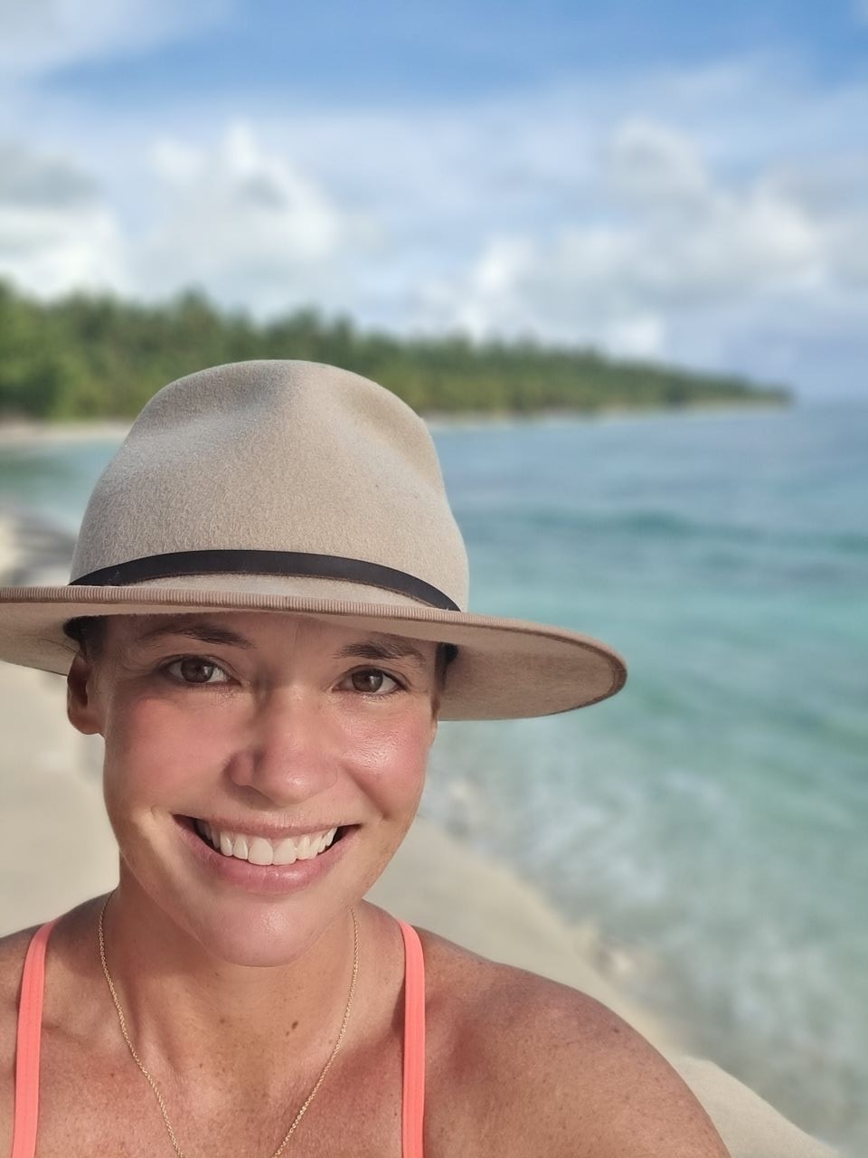 A woman wearing a brimmed hat on a pristine white sandy beach smiles at the camera.
