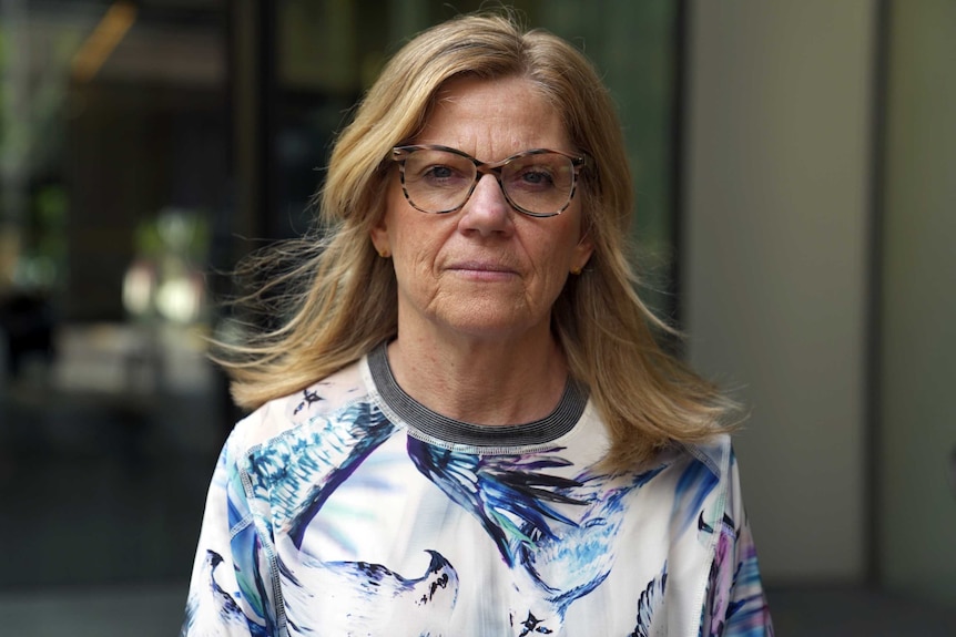 Women with fair hair wearing glasses and colourful top stands looking serious with blurred office building behind