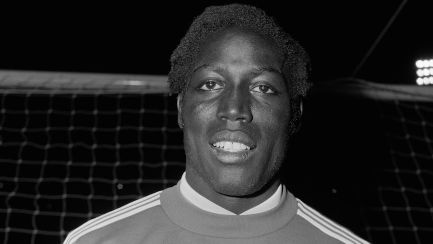A black and white photo of footballer Jean-Pierre Adams standing in front of a goal wearing a Nimes Olympique Adidas tracksuit.