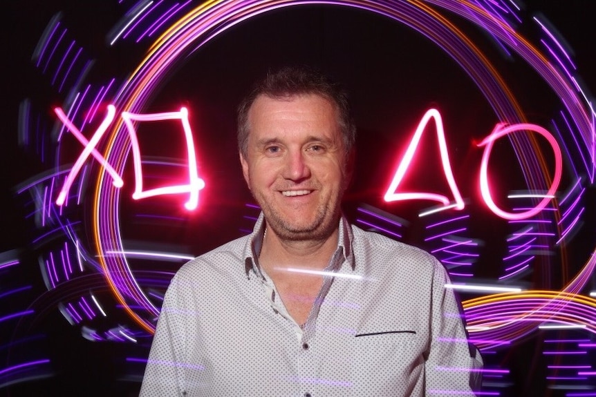 Ross Symons smiling in front of an illuminated sign with playstation symbols. 