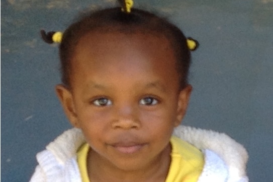 Two-year-old toddler Safa Annour, who is wearing a yellow top, white jumper and jeans, smiles into the camera.