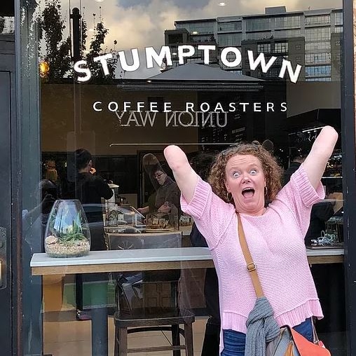 A woman poses in front of a shop called 'Stumptown coffee', gleefully raising her arms, which are both amputed at the elbow.