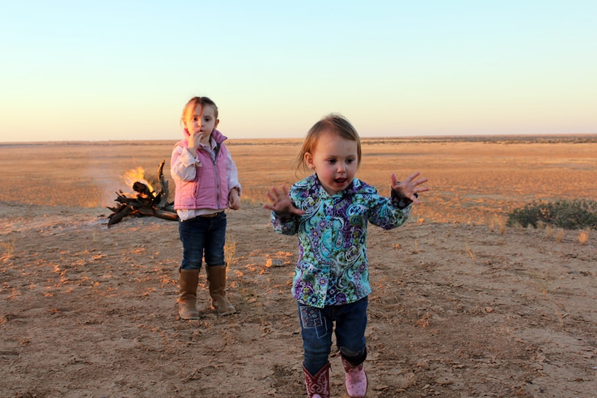 Two young girls run towards the camera in the Simpson Desert at sunset, with a camp fire burning in the distance.