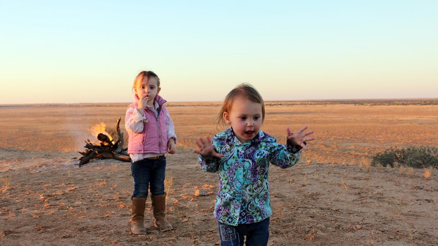 Two young girls run towards the camera in the Simpson Desert at sunset, with a camp fire burning in the distance.