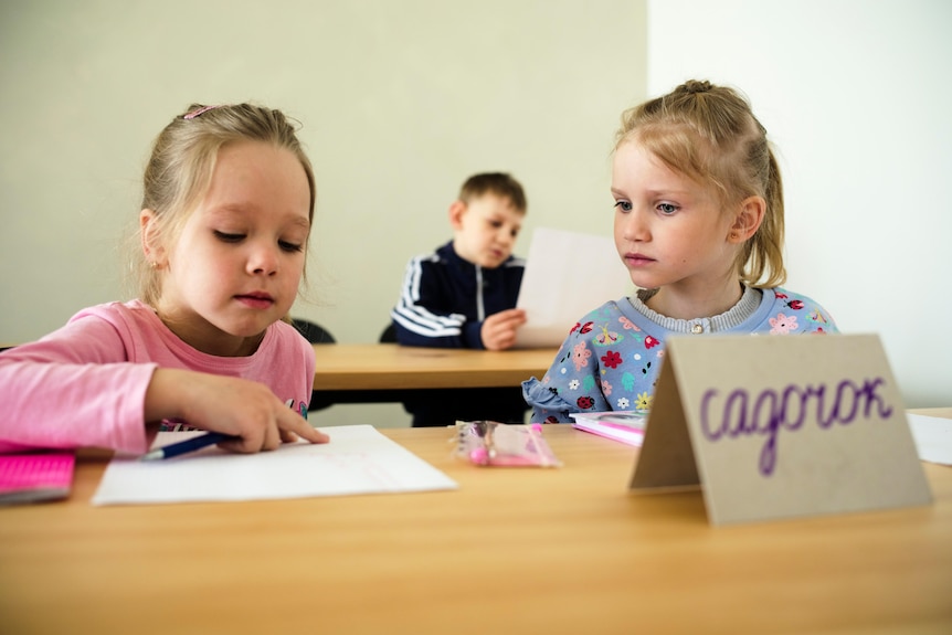 two little girls sit at a classroom desk reading from sheets of paper