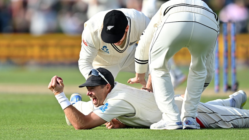 Tim Southee smiles on the ground holding onto a ball with two men hitting his back