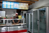 A middle-aged man in a black T-shirt and white cap stands behind an empty food counter, beside empty fridges.