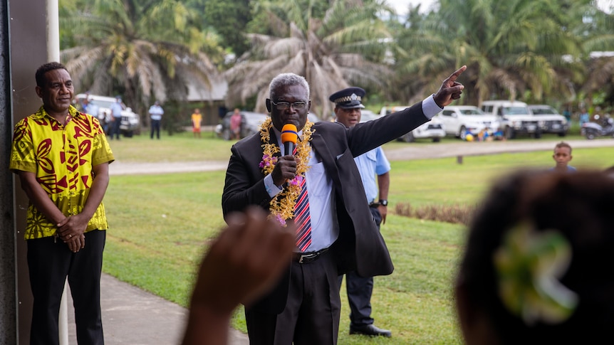 A man wearing a suit and a flower garland around his neck holds a microphone and points upwards.