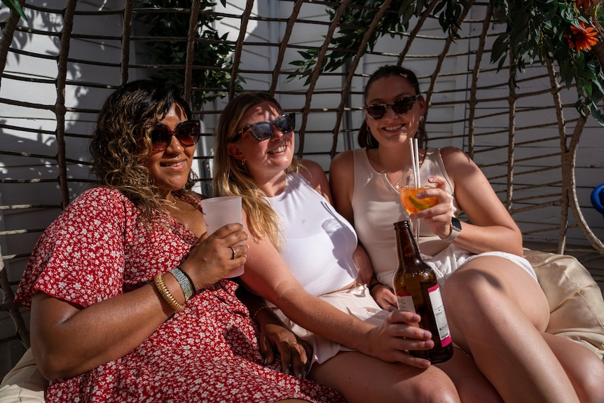 Three women wearing sunglasses hold drinks while sitting in the sunshine.