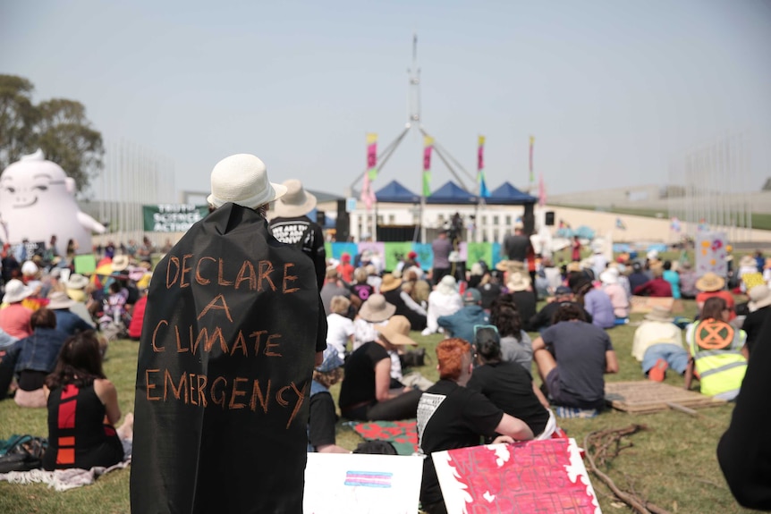 A campaigner is wearing a black cape with "declare a climate emergency" written on it, in front of Parliament House in Canberra.
