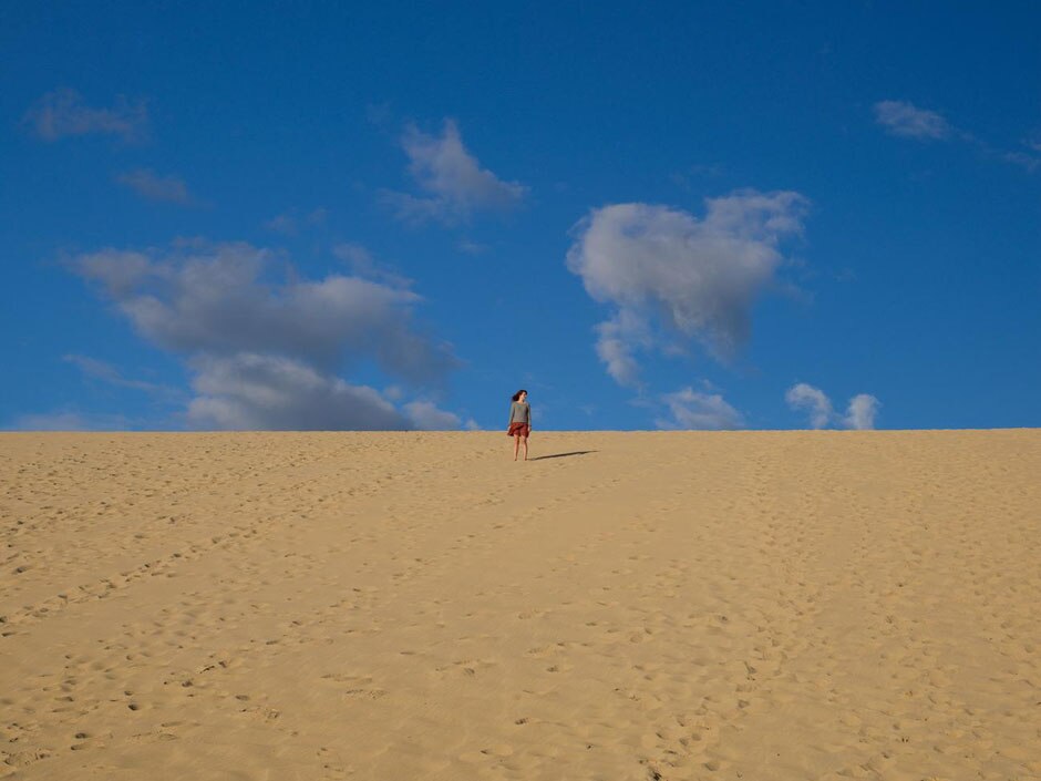 Long shot of a woman standing on a sand hill that's covered in footprints. There's a bright, blue sky beyond with a few clouds.