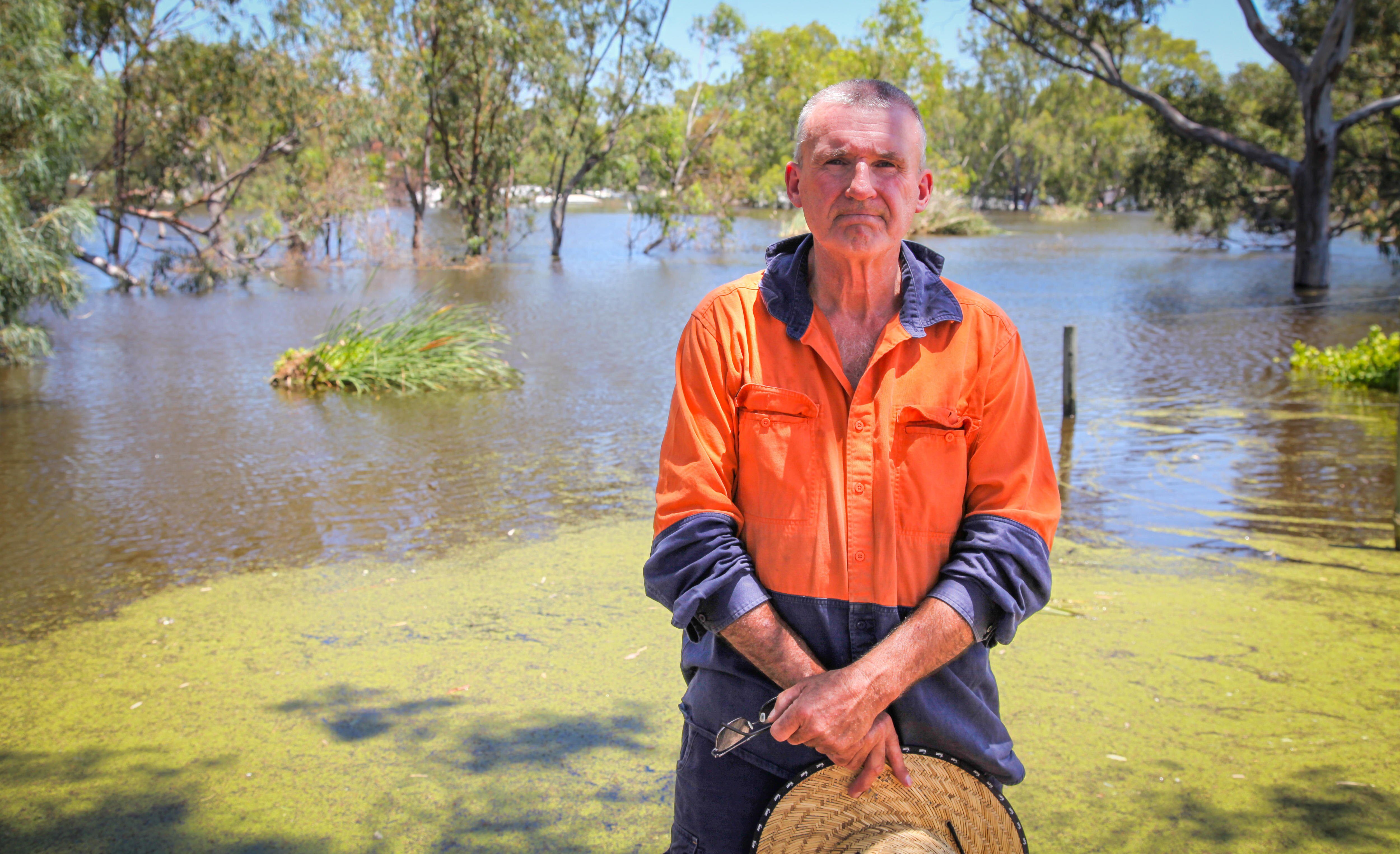 A man in an orange and blue shirt stands holding a hat, behind him is the river