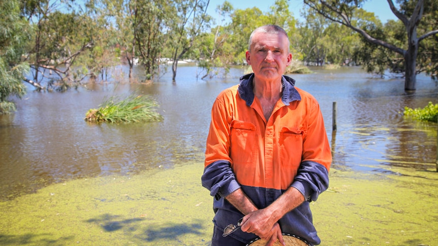 A man in an orange and blue shirt stands holding a hat, behind him is the river