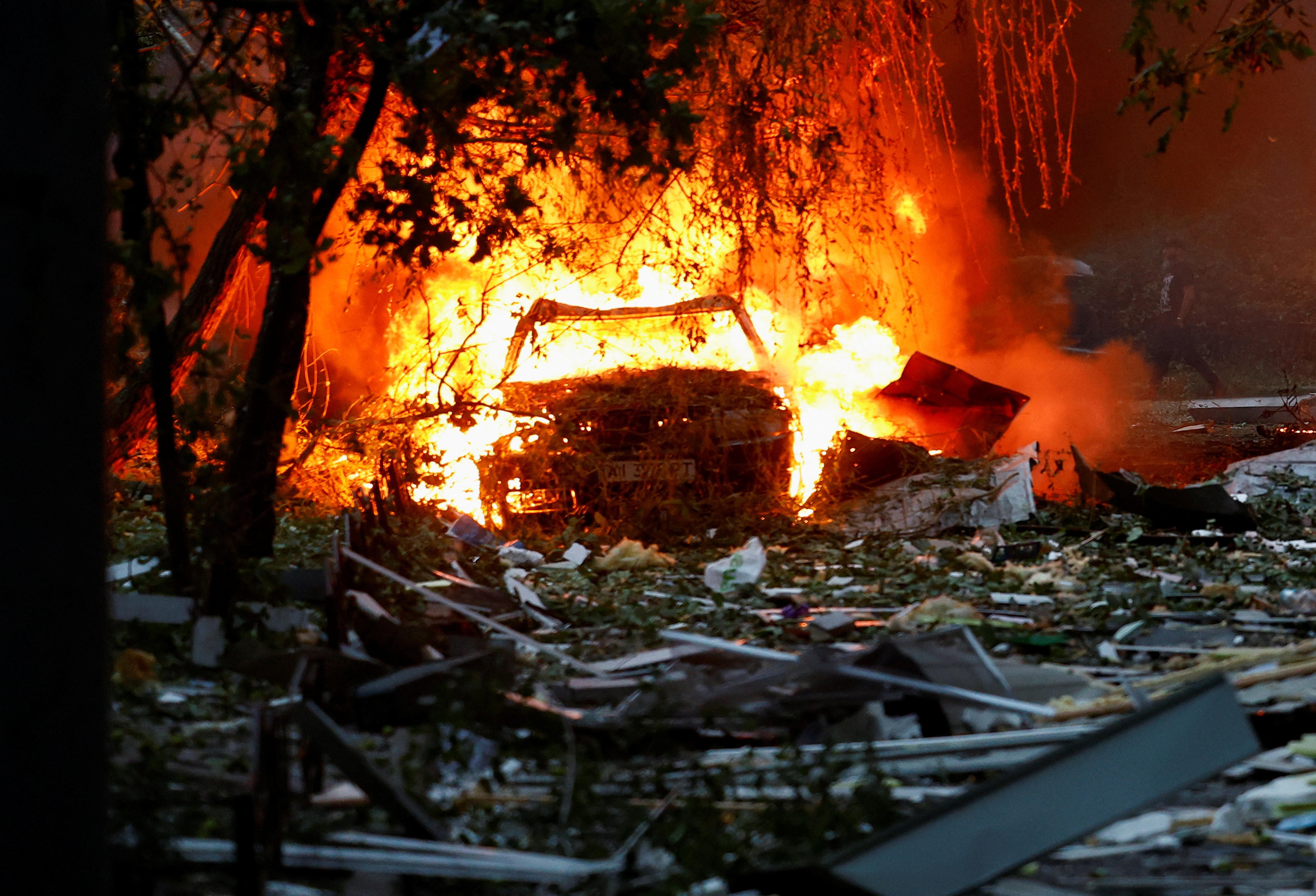 A charred wreckage of a car illuminated in bright orange and red flames on piles of building rubble