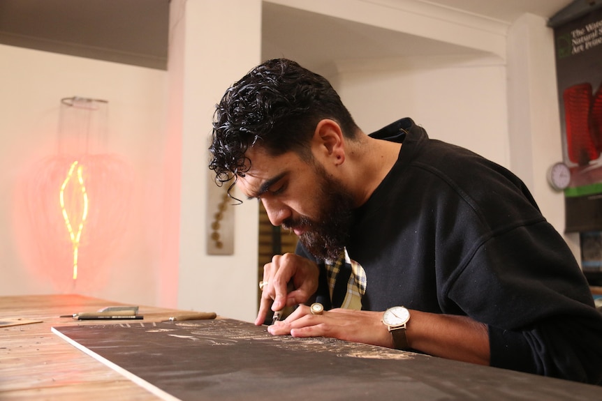 A man bends over a plank of wood, carving.