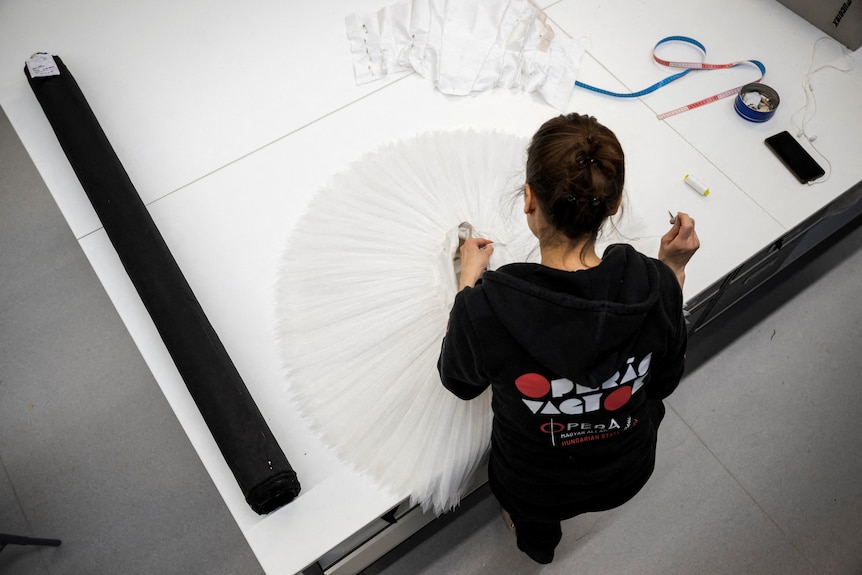 A seamstress works on a white ballet dress that lays on a table in front of her.