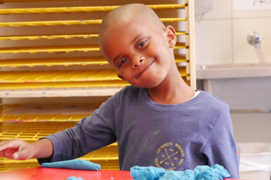 A young boy in a blue shirt with a shaved head smiles for a photo sitting at a table playing with playdough.