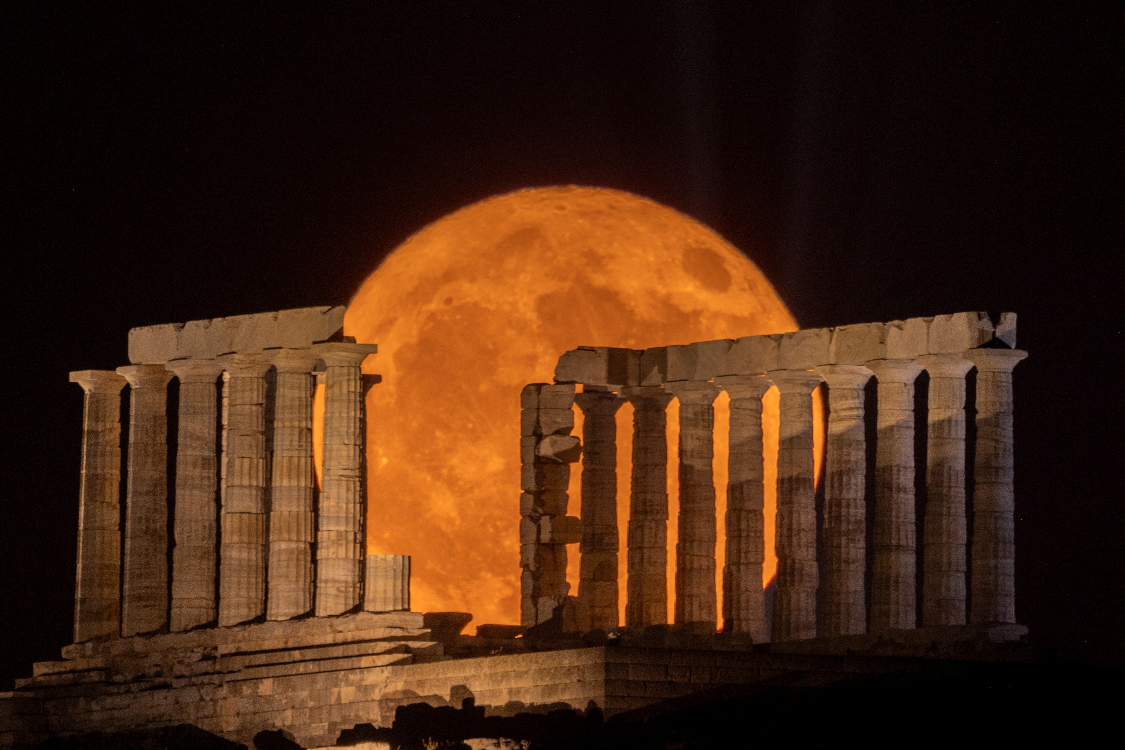 A huge orange moon rises behind a ruined Grecian temple with two rows of damaged stone columns.