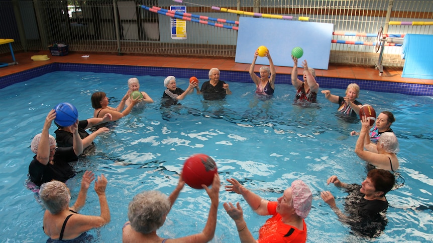 A group of older people stand in a circle chest-deep in water in an indoor pool, passing balls to one another.
