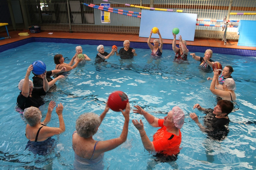 A group of older people stand in a circle chest-deep in water in an indoor pool, passing balls to one another.