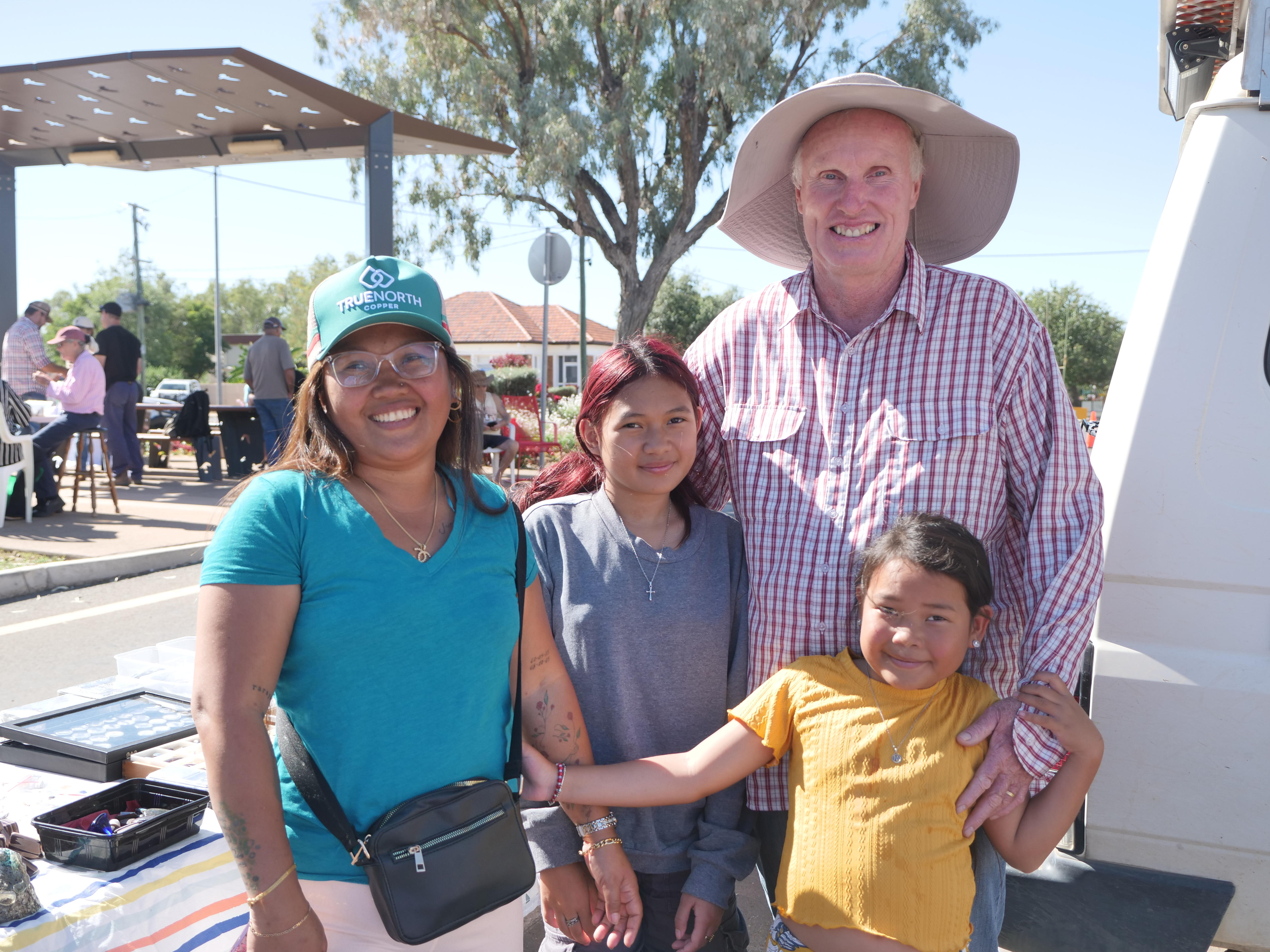 A smiling woman, man and two young girls standing near an open-air market stall.