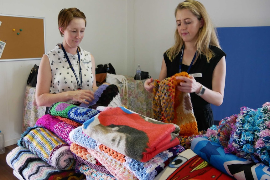 Two young women wearing work clothes fold hand knitted blankets in a hall. They're concentrating on their work.