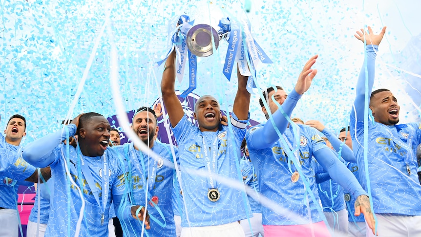 Manchester City players lift a trophy with blue ticker tape behind them