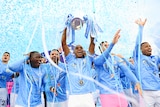 Manchester City players lift a trophy with blue ticker tape behind them