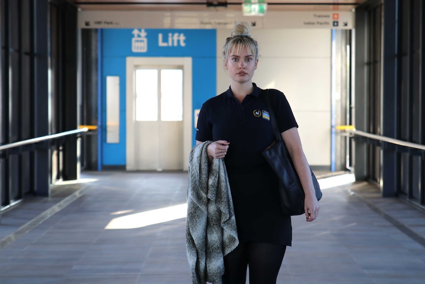 Wanita holds her jacket and bag, and stands in a corridor with signs for train station platforms behind her.