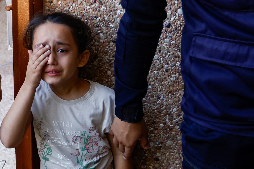 A young girl wipes a tear while she cries, with an adult standing by her