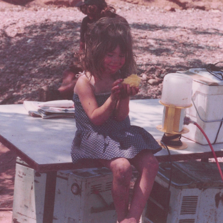 Aticia at four, sitting on a table in the red dirt, hair in a knot, devouring a mango.