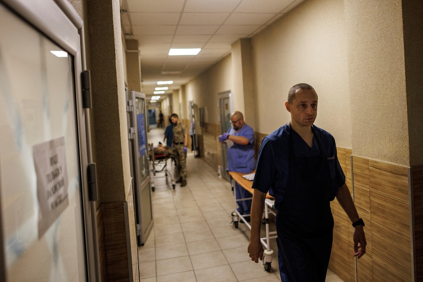 A doctor in blue scrubs walks down a hall as other staff and military stand against wall behind