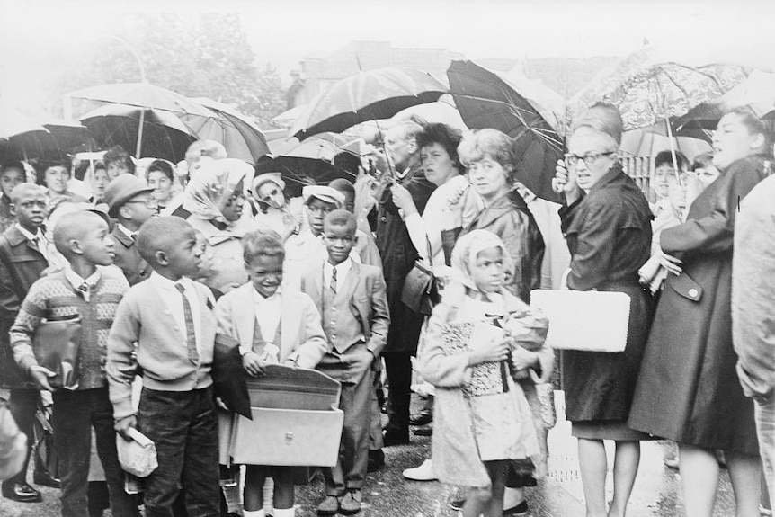 A black and white photo of young black children walking past a line of white women with umbrellas