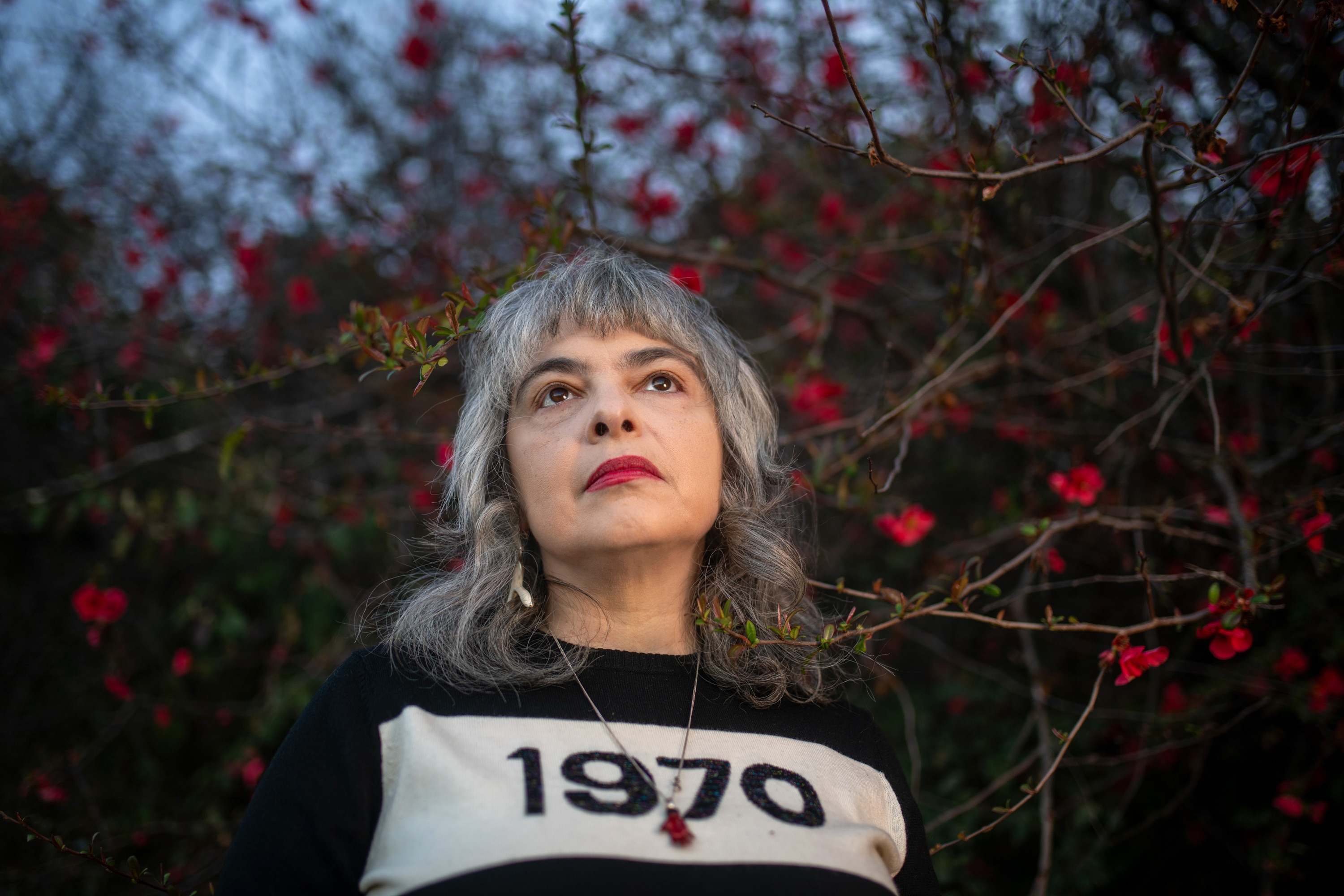 A woman with red lipstick against a hedge of red flowers and spikey twisted twigs.
